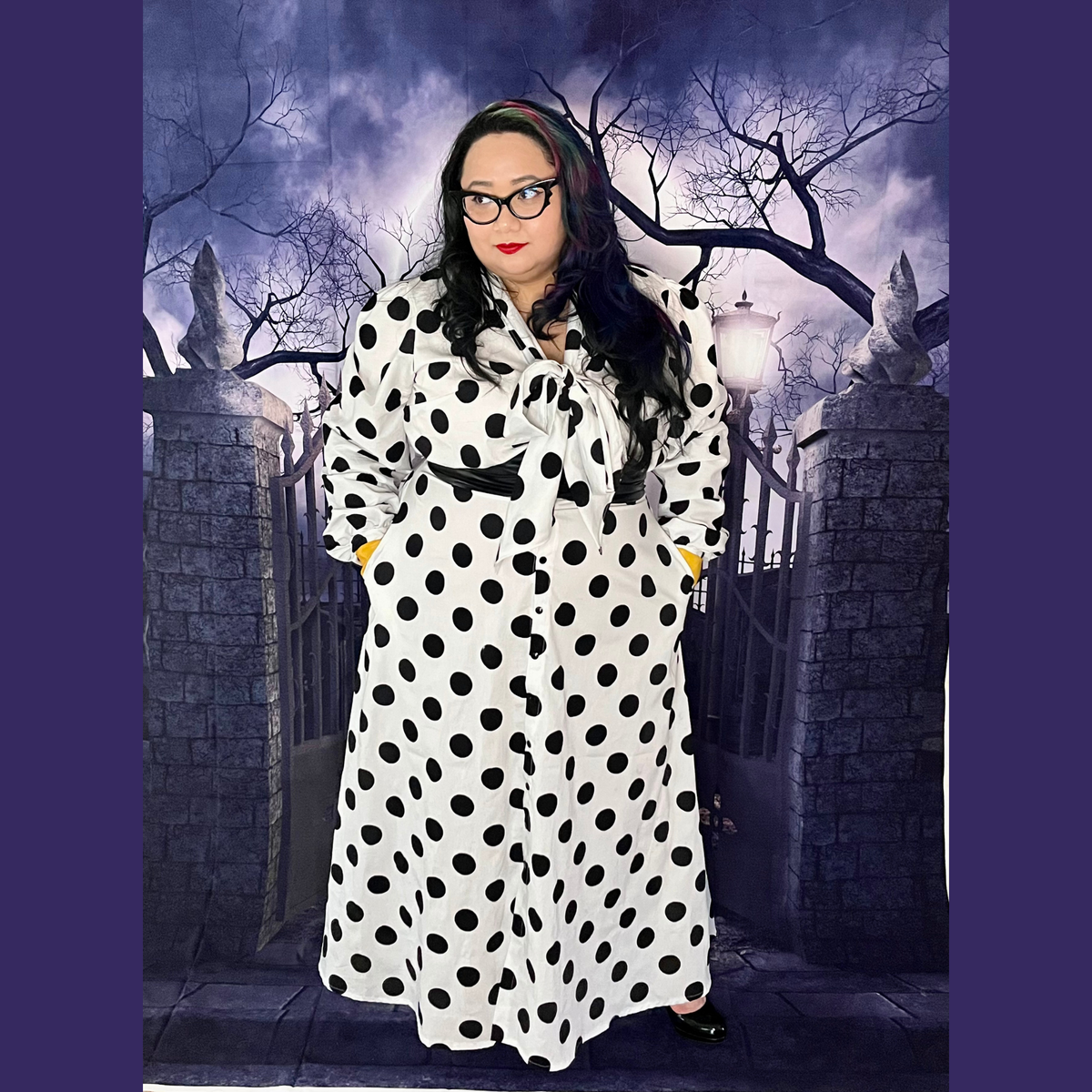A person wearing a long-sleeve white polka-dot maxi dress with a tie neckline, styled with bright yellow gloves and glasses, standing in front of a spooky gothic gate backdrop.