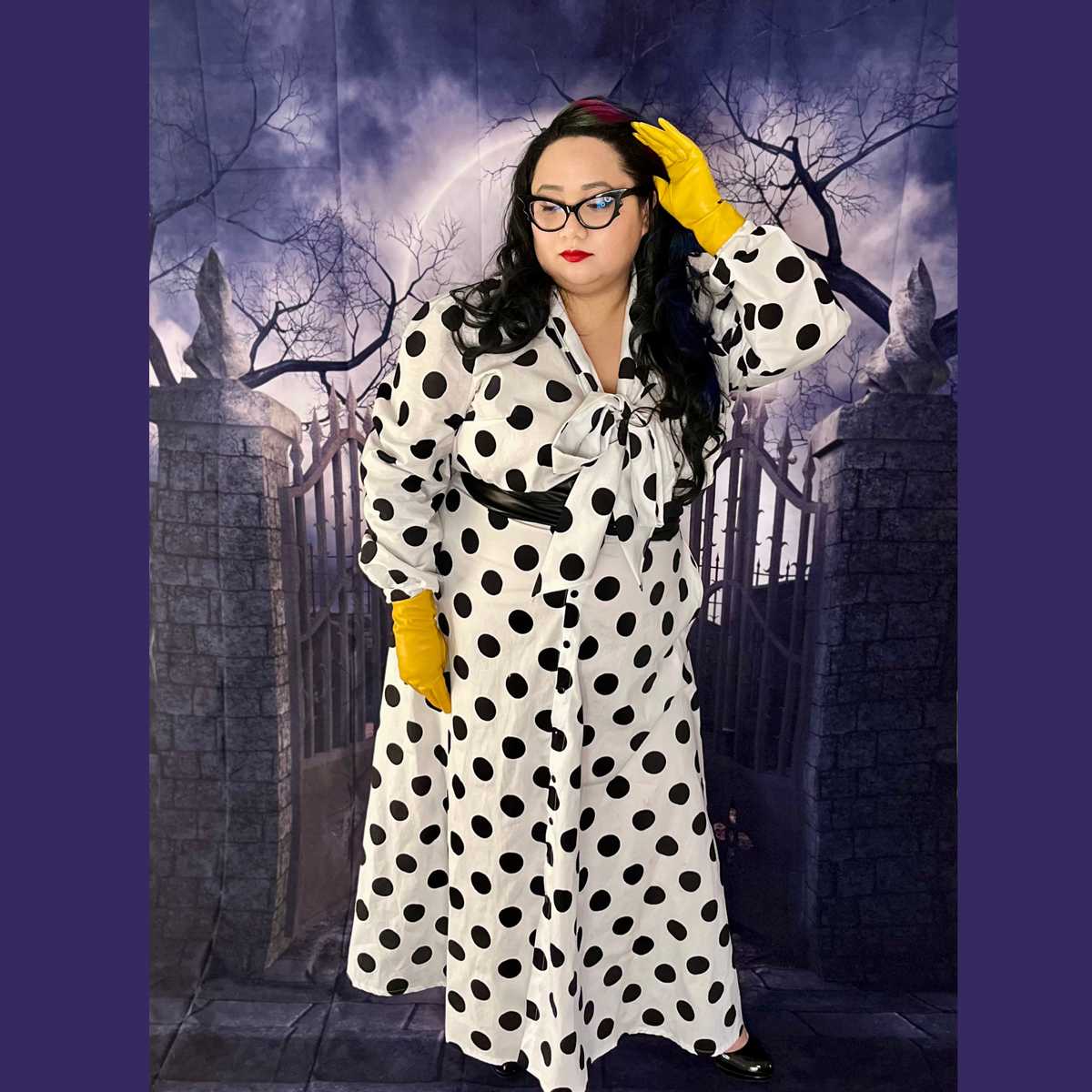 A woman modeling a white long-sleeve dress with large black polka dots, styled with bright yellow gloves and glasses, standing in front of a spooky gothic gate backdrop.