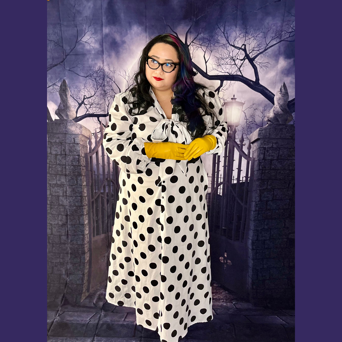 A person wearing a long-sleeve white polka-dot maxi dress with a tie neckline, styled with bright yellow gloves and glasses, standing in front of a spooky gothic gate backdrop.