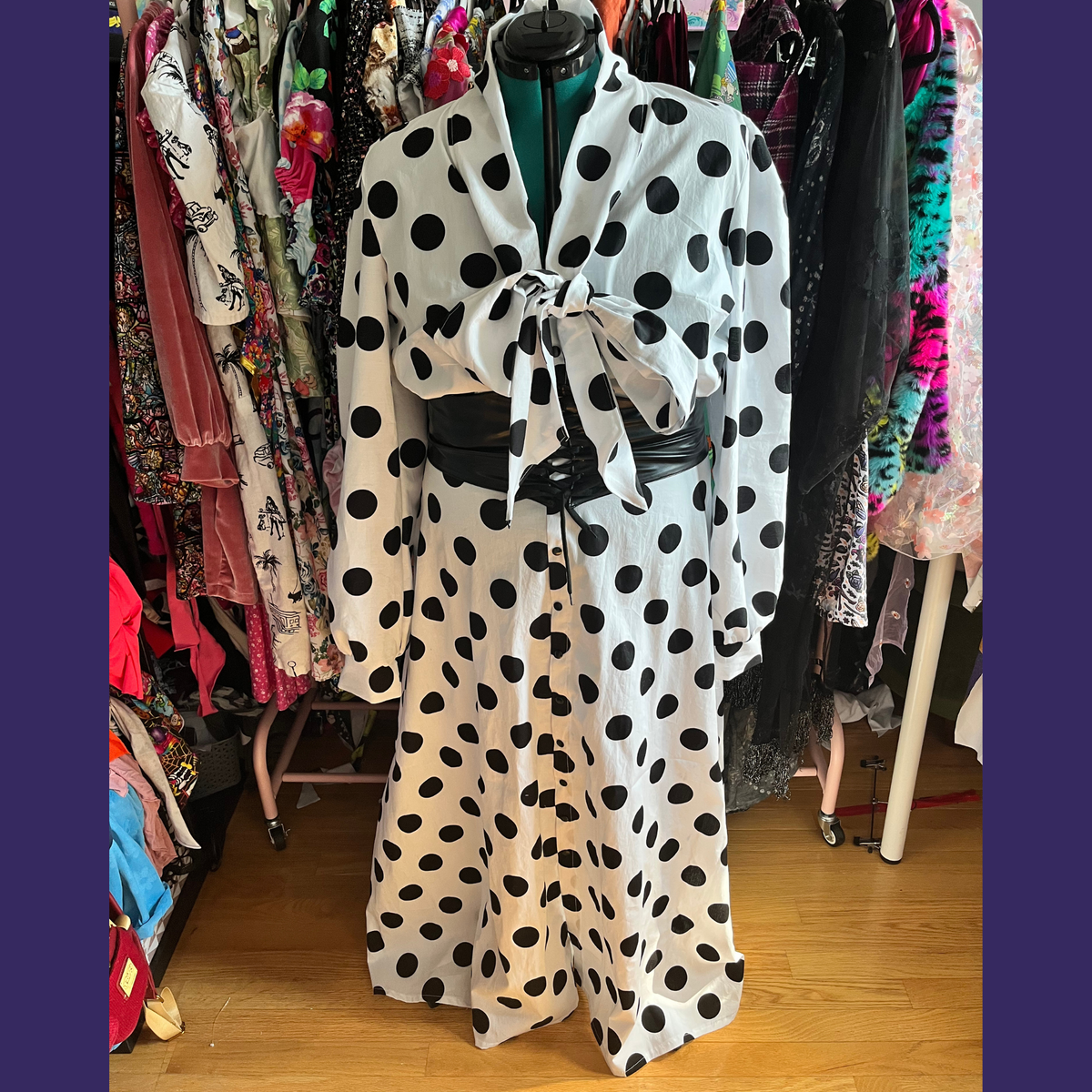 A long-sleeve white blouse or dress with large black polka dots, displayed on a mannequin torso in a clothing store. The garment has a tie at the collar and a black belt at the waist.