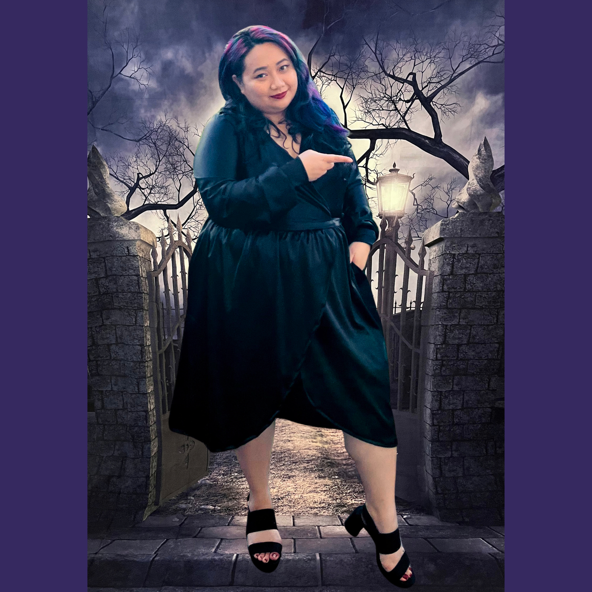 A woman modeling a black wrap-style dress in a gothic cemetery setting with stone gates and bare trees. The dress has long sleeves, a V-neckline, and a gathered waist.