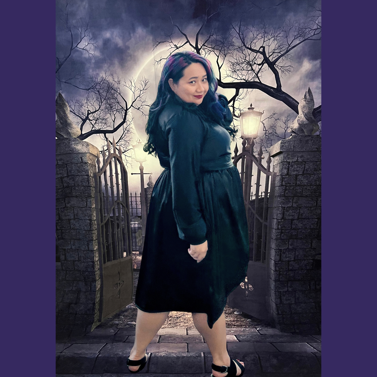 A woman modeling a black wrap-style dress in a gothic cemetery setting with stone gates and bare trees. The dress has long sleeves, a V-neckline, and a gathered waist.