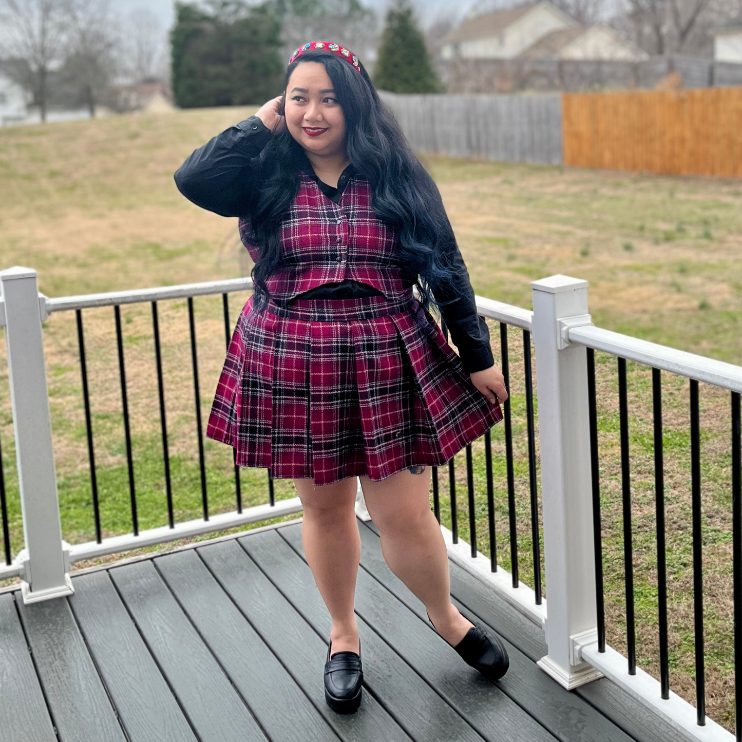 Person wearing a red plaid dress and black jacket on a deck with grass and fence in the background