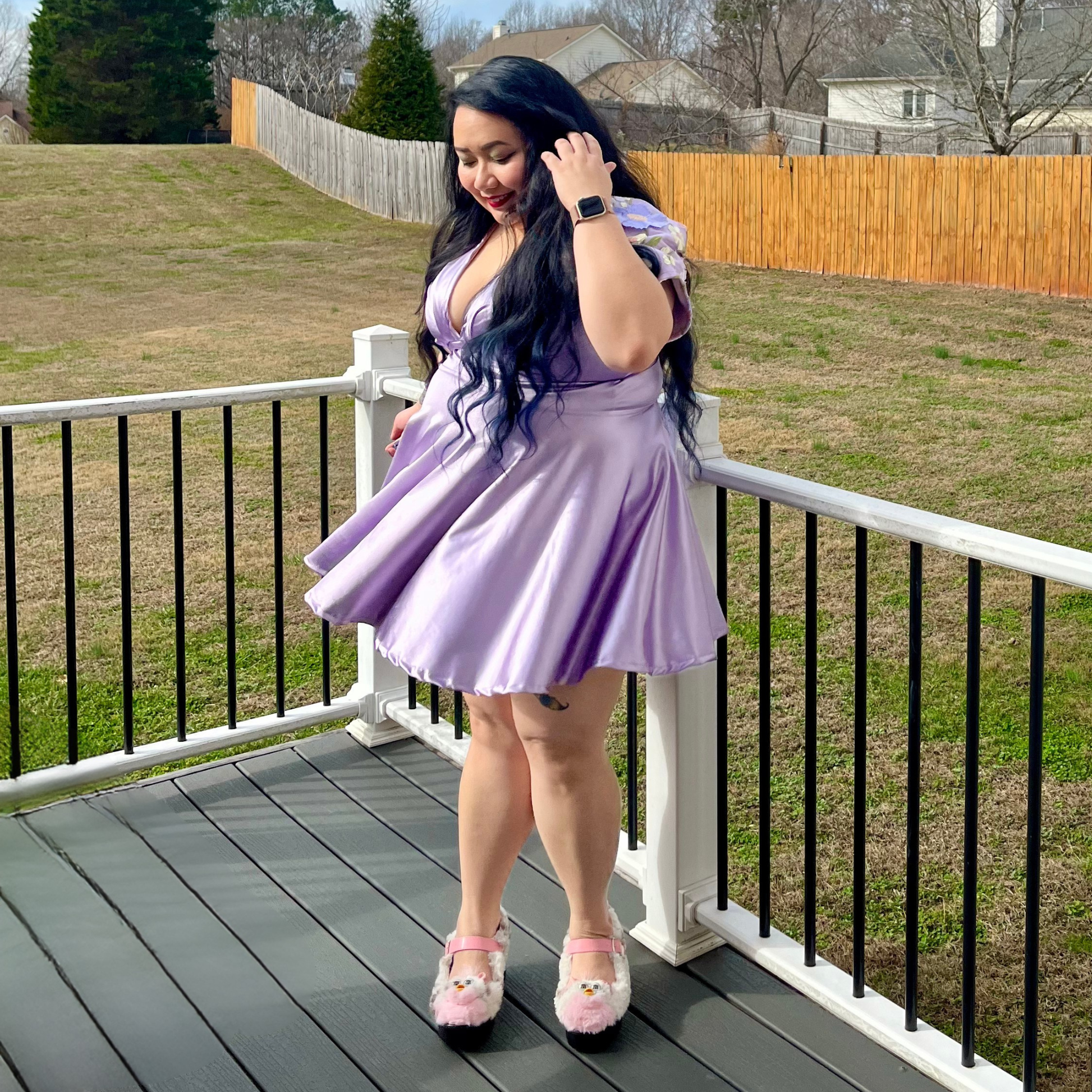 Woman in a purple dress standing on a deck with a grassy area and wooden fence in the background.