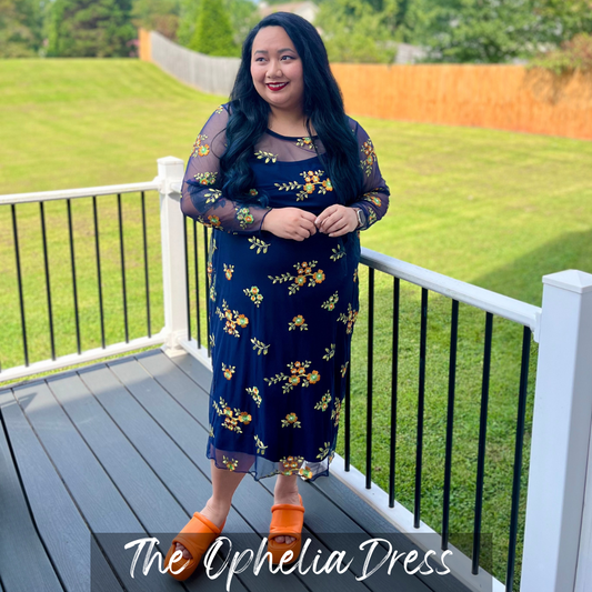 Woman wearing a navy blue dress with floral patterns on a deck with green grass and trees in the background.