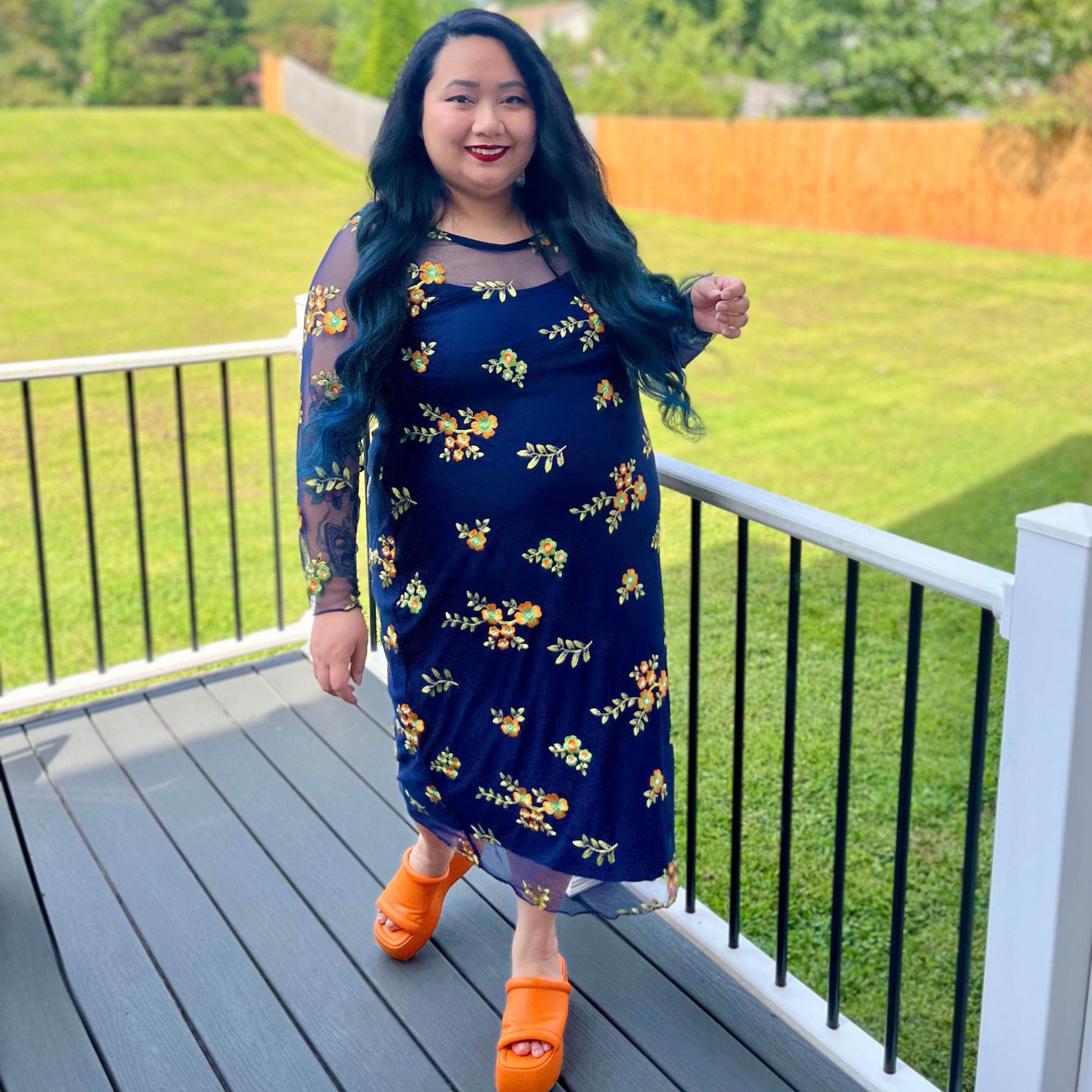 Woman in a blue floral dress standing on a deck with green grass and trees in the background