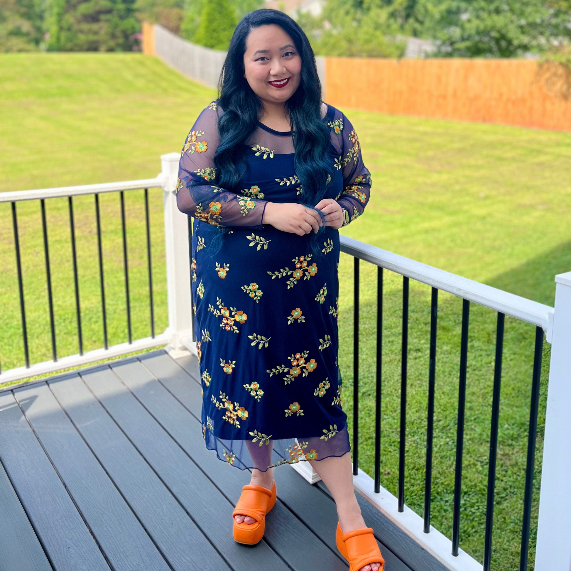 Woman in a blue floral dress with orange shoes standing on a deck with green grass and trees in the background