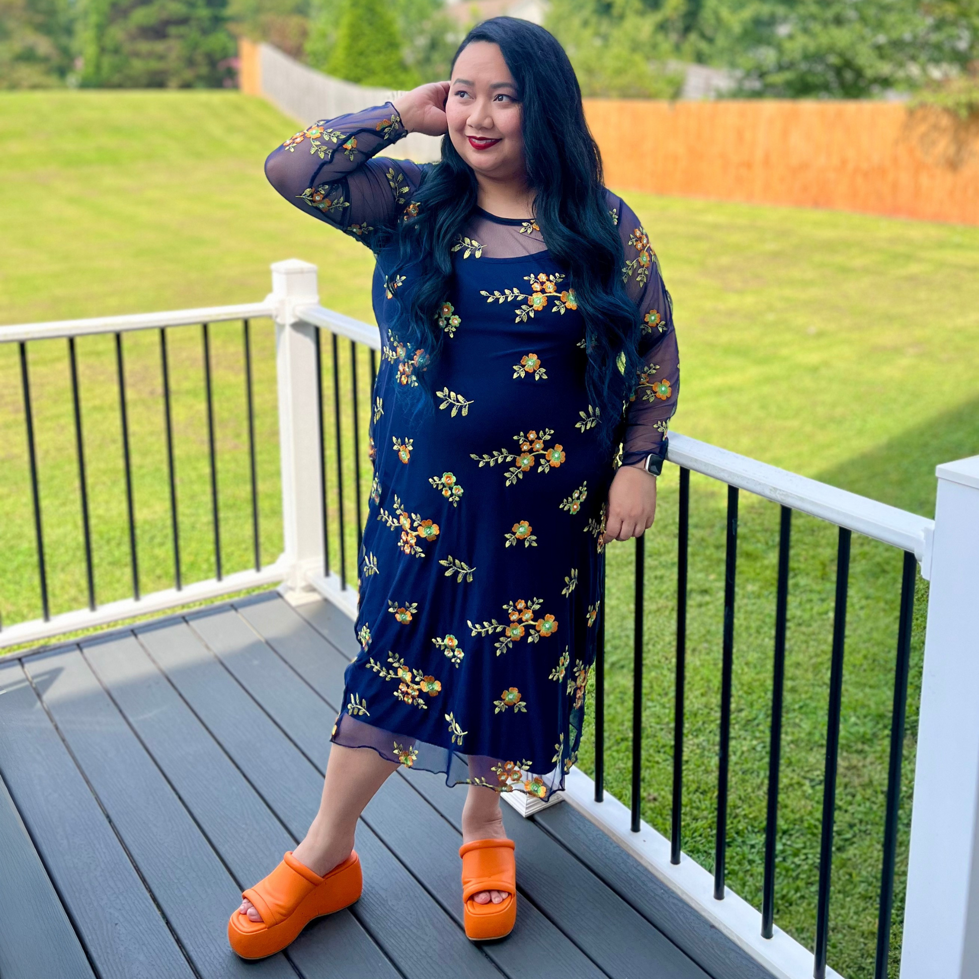 Woman in a blue floral dress with orange sandals standing on a deck.