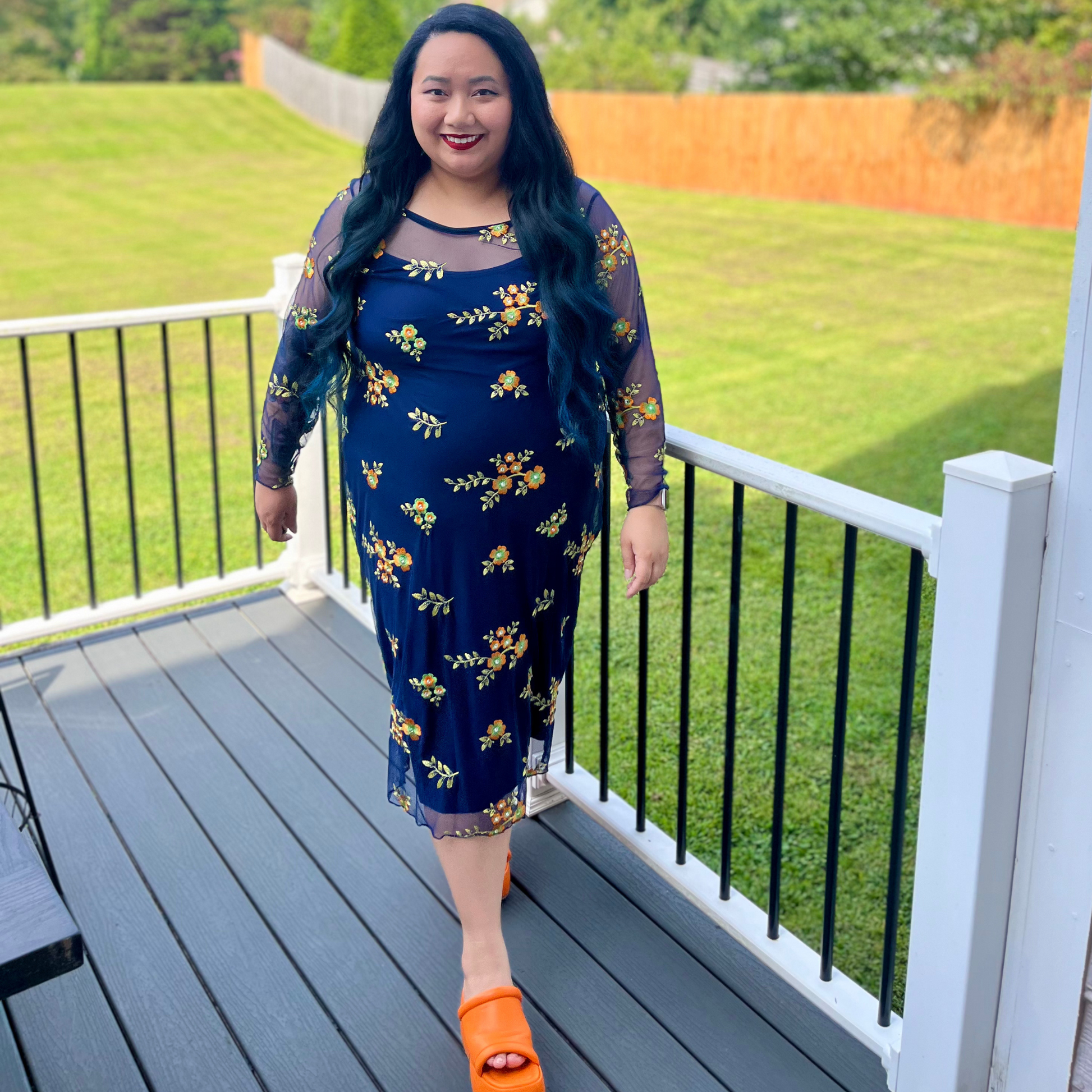 Woman in a blue floral dress standing on a deck with green grass and trees in the background